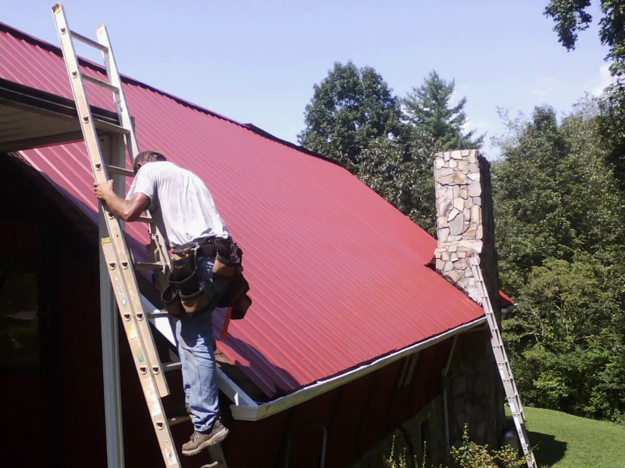 Roofer on a ladder beside a red metal roof and stone chimney — Soffit & Fascia Repair by Roofing Brothers in Oaks